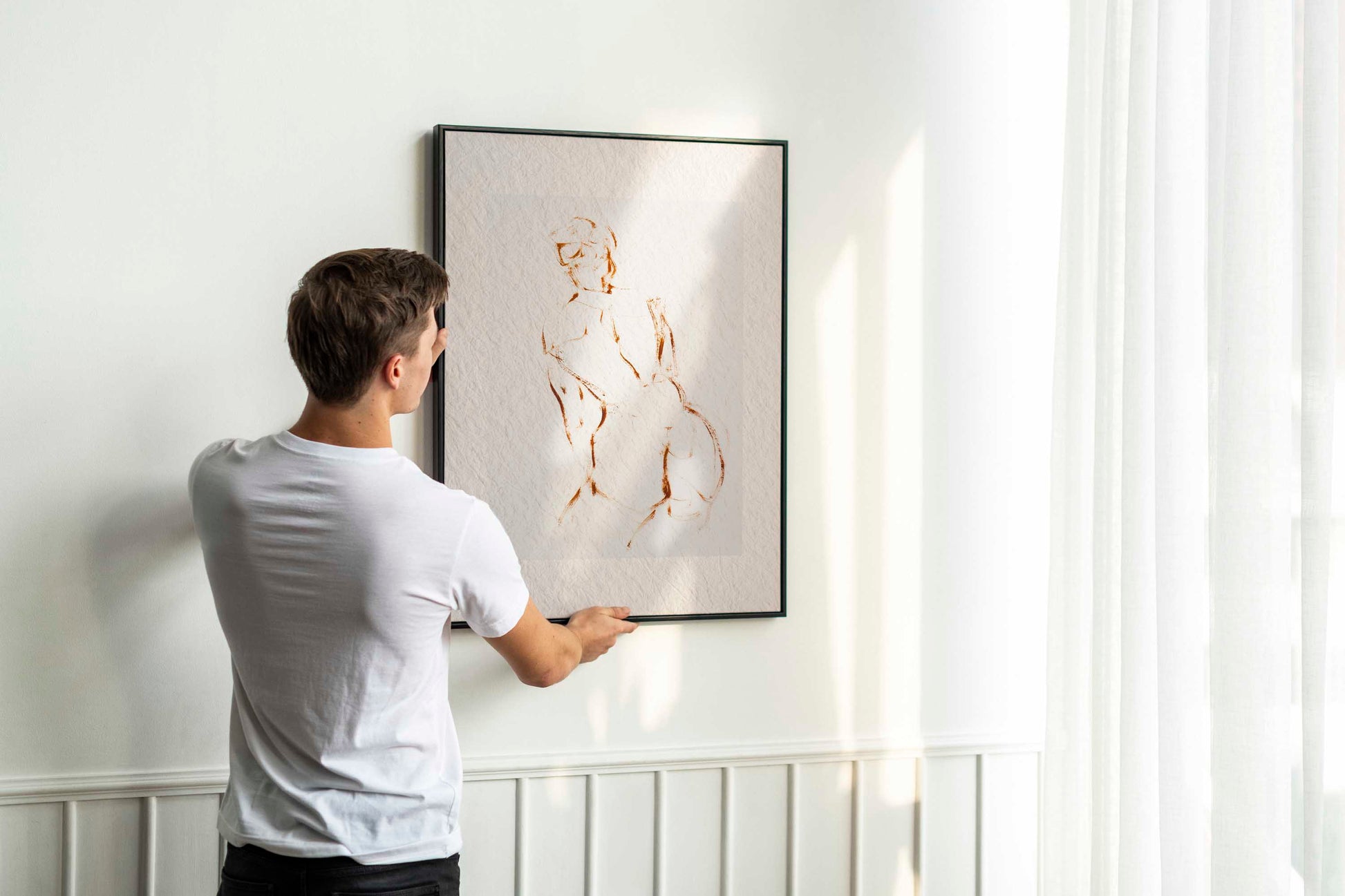 Person holding up framed A Shape I Almost Knew fine art print by Bogdan Jensen on a wall, showing the minimal brown line drawing in a bright interior