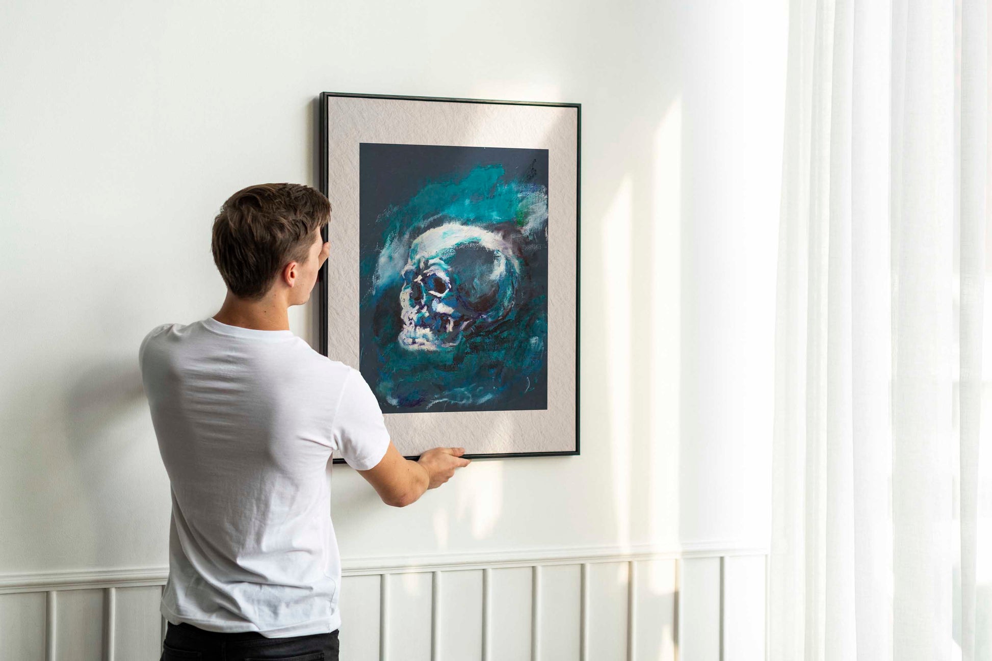Person holding up framed Blue Remains fine art print by Bogdan Jensen on a wall, showing the blue skull study in a bright interior