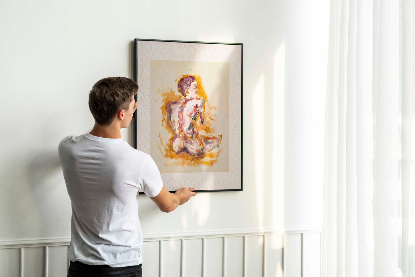Person holding up framed Felt through Gold fine art print by Bogdan Jensen on a wall, showing the seated figure painting in warm gold and lilac tones in a bright interior