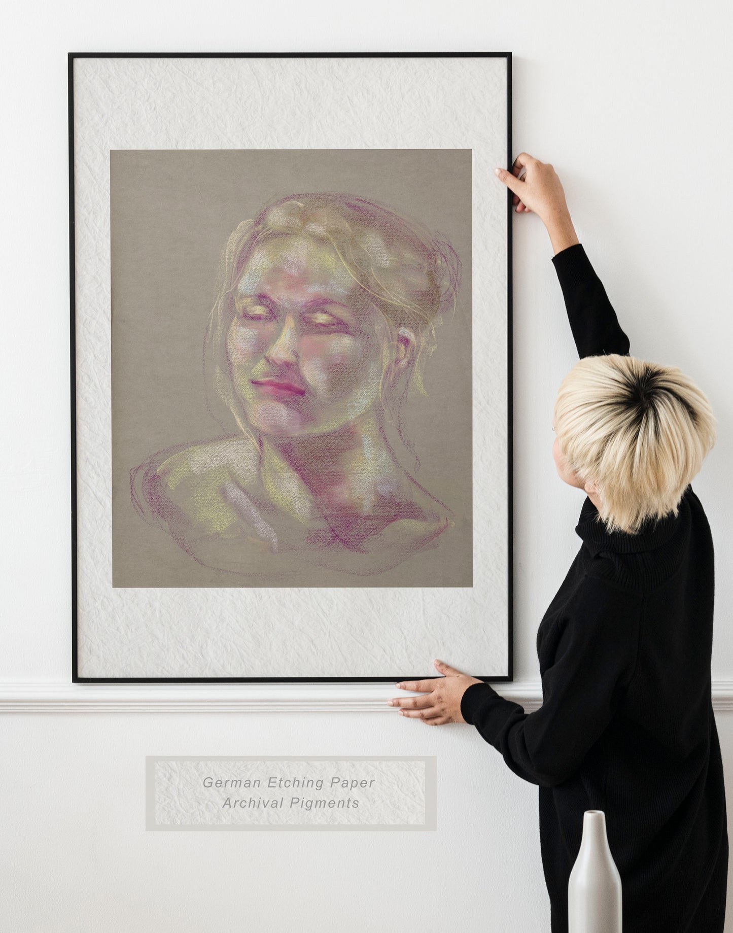 Woman holding framed Soft Glow fine art print by Bogdan Jensen on a wall, showing a soft pastel portrait in pale pink, yellow, and violet tones