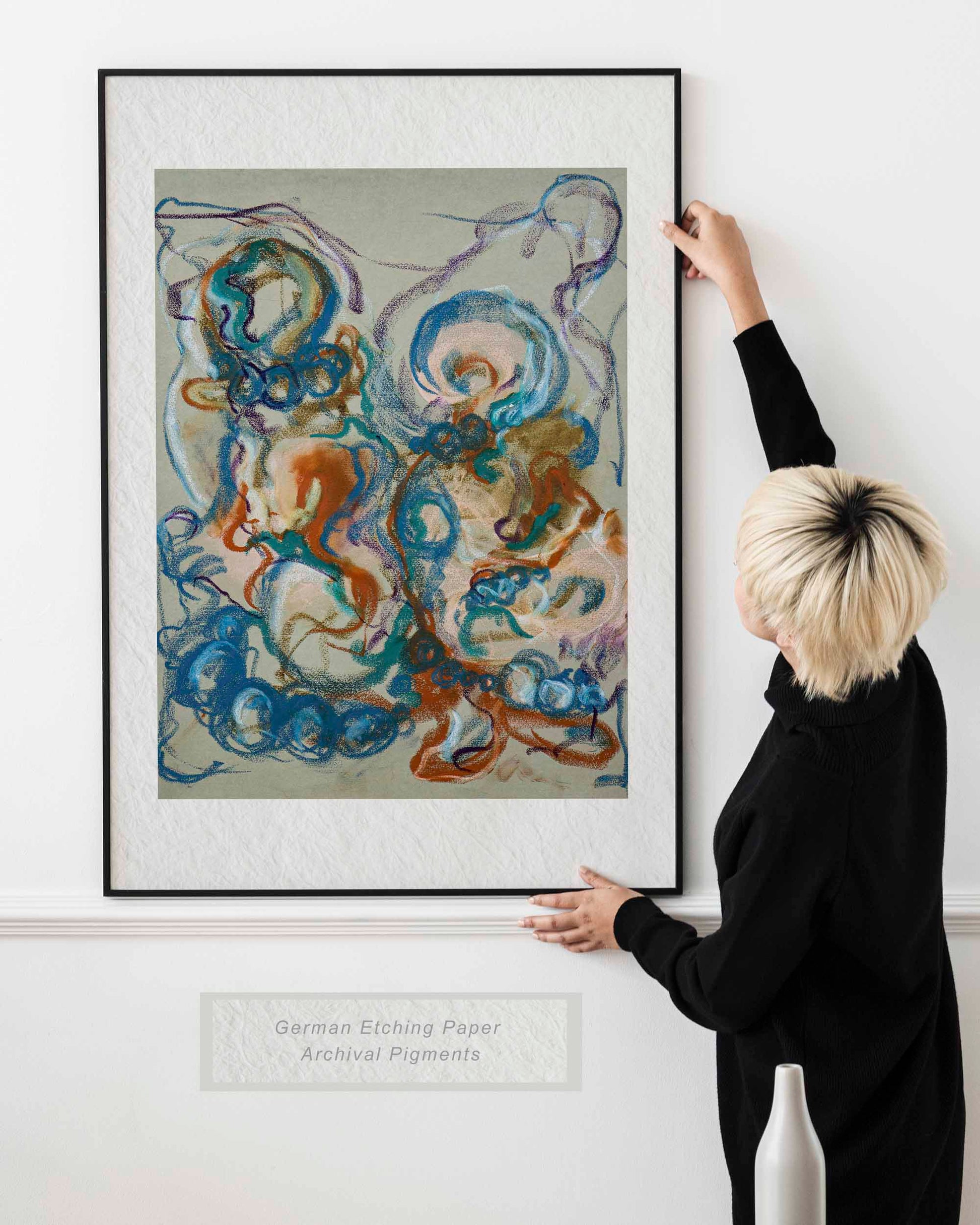 Woman holding framed Together in the Garlands fine art print by Bogdan Jensen on a wall, showing abstract figurative forms in blue, ochre, teal, and violet