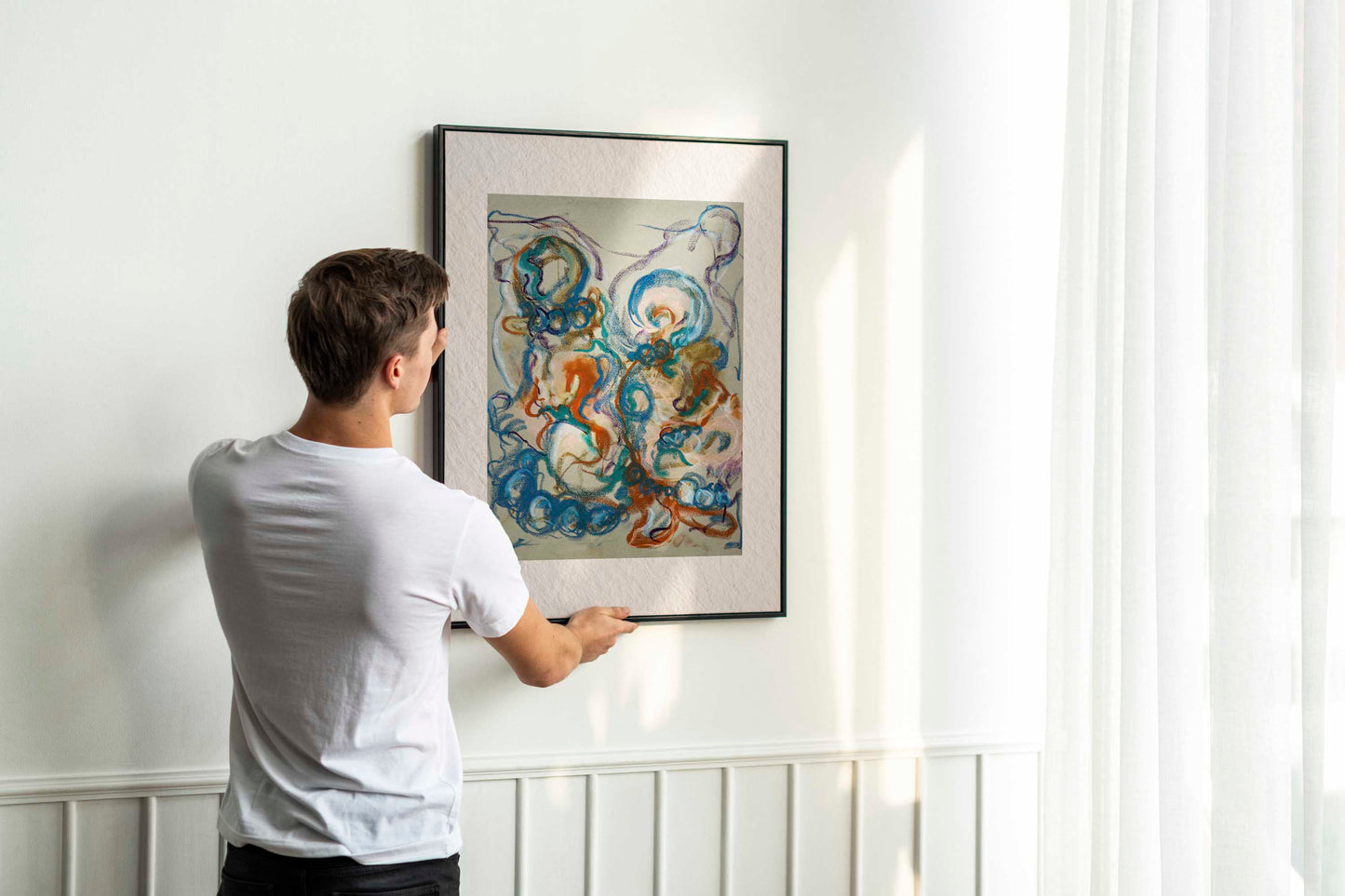 Person holding up framed Together in the Garlands fine art print by Bogdan Jensen on a wall, showing the colourful abstract figurative drawing in a bright interior