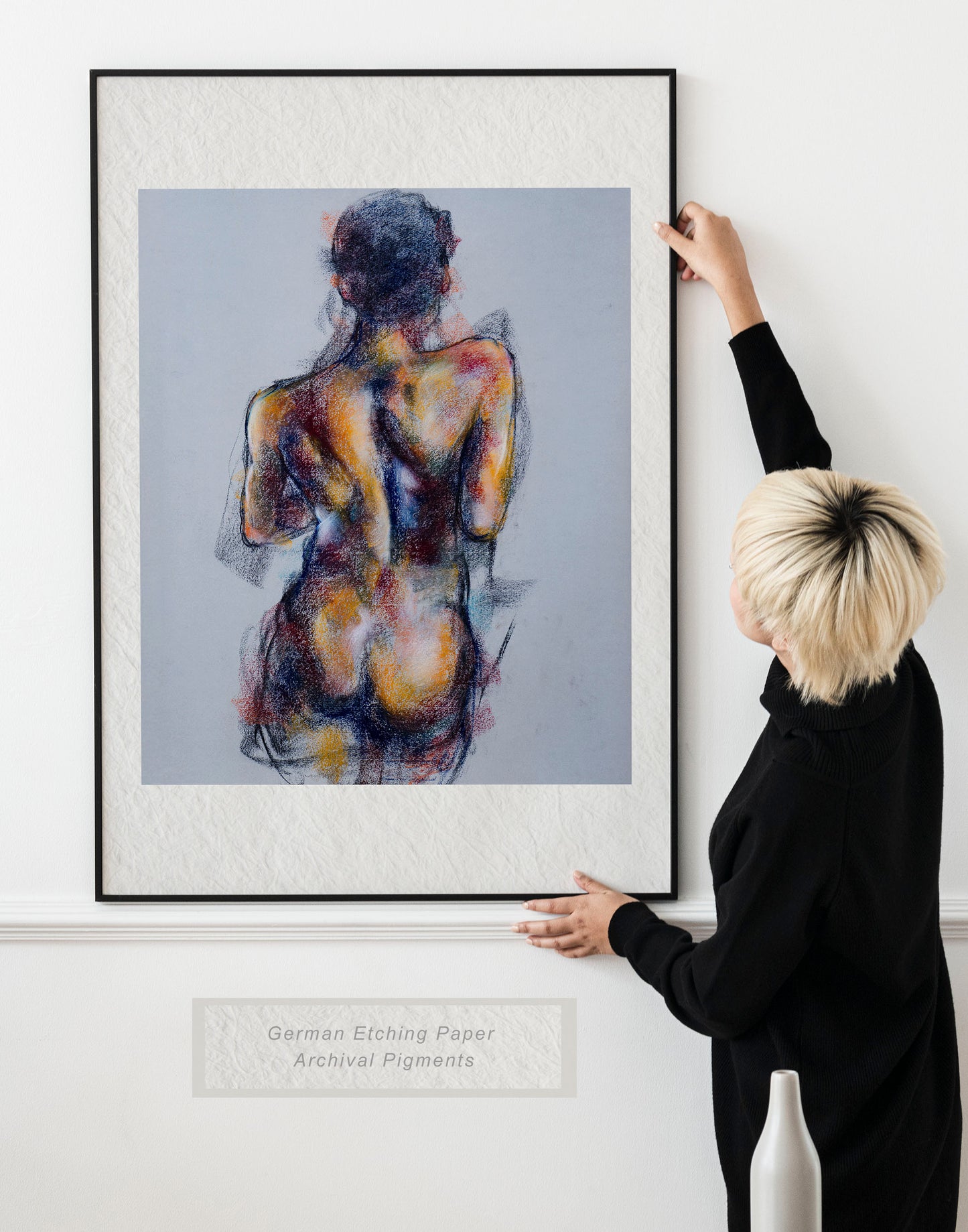 Woman holding framed Where the Dance Lives fine art print by Bogdan Jensen on a wall, showing a soft pastel figure study of a dancer’s back in a bright interior